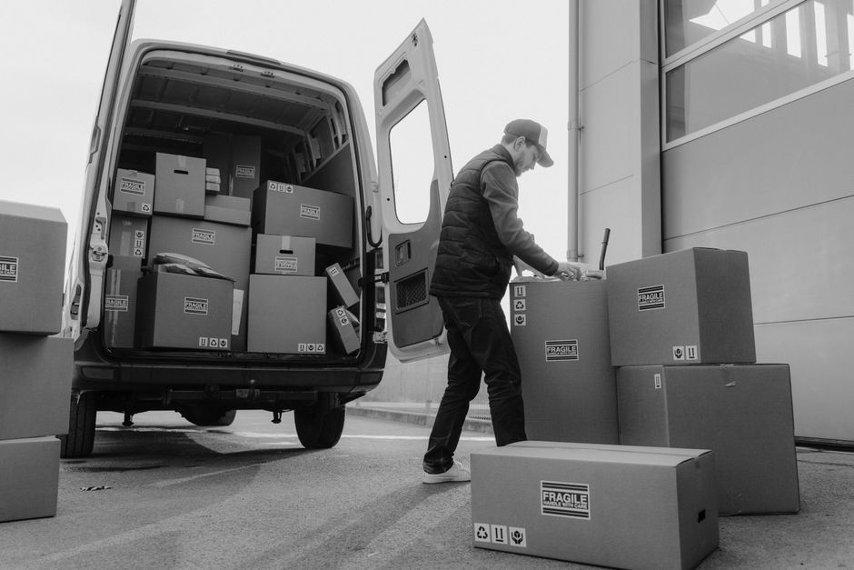 A man wearing a dark uniform is seen loading a large cardboard box into a van, which is being used for house removals. The box is sealed with packing tape and has printed symbols indicating handling i