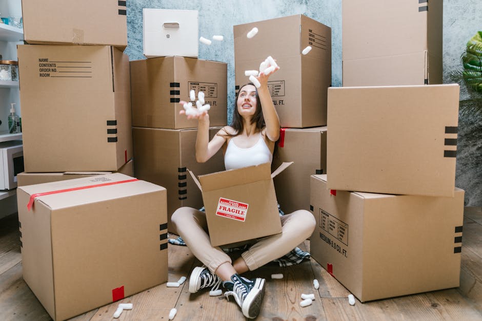 A young woman with long dark hair, sitting cross-legged on a wooden floor indoors, surrounded by numerous large cardboard boxes of varying sizes, some sealed with red tape and others open. She is wear