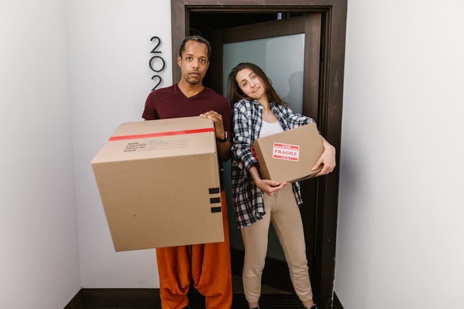 A young woman with long dark hair, sitting cross-legged on a wooden floor indoors, surrounded by numerous large cardboard boxes of varying sizes, some sealed with red tape and others open. She is wear