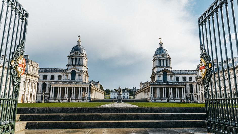 A view through an open wrought iron gate showing two white, neoclassical buildings with domed roofs and columns, set on a well-maintained lawn with steps leading up to them. The sky is partly cloudy w