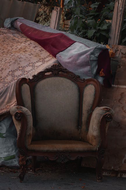 A pink, upholstered sofa with a tufted backrest and armrests, showing signs of damage with torn fabric and stuffing exposed on the left side. The sofa is placed outdoors on a paved pavement adjacent t