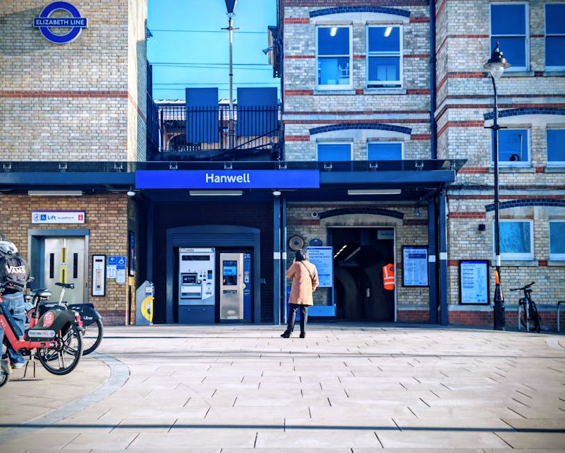 A person standing outside the Hanwell station entrance, which features a brick facade with decorative red brick accents and large windows. The station sign is displayed on a black awning above the doorway. In front of the entrance, there are several electric bike and scooter rental stations, with some bikes parked nearby. To the left of the entrance is a small kiosk or ticket machine area, and to the right, a dark archway leading into the station. A street lamp is mounted on the building's corner, and an orange high-visibility jacket is visible on a staff member or worker near the door. The pavement in front of the station is clean with a smooth, tiled surface, providing an unobstructed area suitable for loading and unloading during a home relocation or furniture transport process, supporting the services offered by Man and Van Woolwich. The scene is well-lit with natural daylight, indicating daytime.