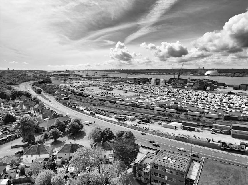 A black-and-white aerial photograph capturing a large industrial port area with numerous shipping containers arranged in rows, multiple cranes, and warehouse buildings. In the foreground, there is a residential neighbourhood with houses, trees, and streets. The image also shows a major road with several vehicles moving along it, separating the residential area from the port. The sky above features scattered clouds and a mix of darker and lighter cloud formations, indicating a partly cloudy weather. The image provides a broad perspective of the urban landscape, illustrating the scale of the port facilities in contrast to the nearby homes. This scene relates to home relocation and furniture transport logistics, as it visually represents the kind of environment where professional removals services, like those provided by Man and Van Woolwich, operate to assist with packing, moving, and transportation processes near Woolwich Arsenal station.