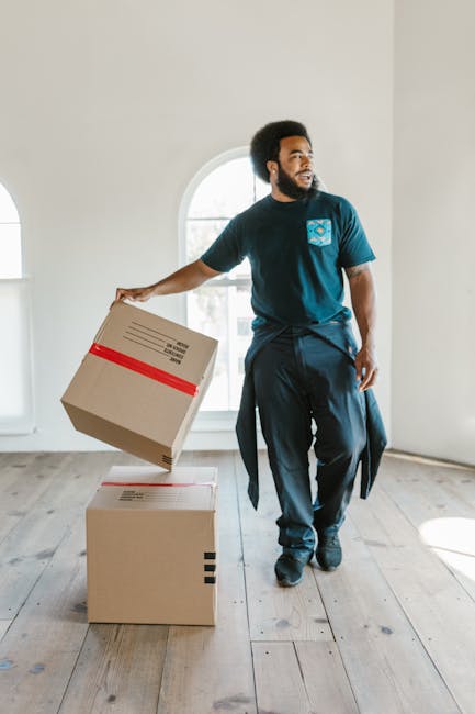 A man wearing a blue t-shirt and dark trousers stands inside a bright, empty room with white walls and a large arched window, holding a cardboard box with red packing tape in his right hand, about to